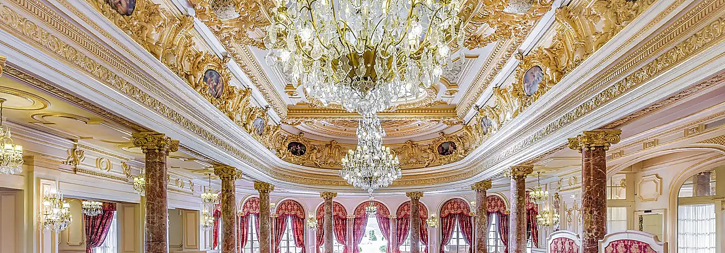 Salón de fiestas con cielorraso dorado, pinturas en medallón, arañas de caño colgantes y columnas de mármol entre cortinados rojos.